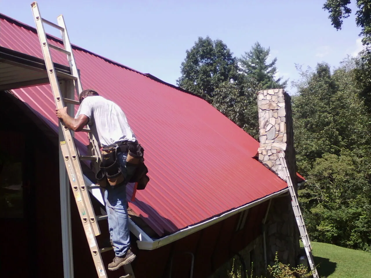 Roofer on a ladder beside a red metal roof and stone chimney — Soffit & Fascia Repair by Roofing Brothers in Crouse